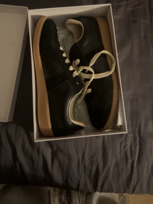 Maison Margiela svarta sneakers - Svarta sneakers från Maison Margiela med klassisk låg siluett, vita snören och beige gummisula. Skorna har en stilren design med paneler i läder och mocka, perfekta för dig som gillar minimalistisk och trendig stil. priset ej hugget i sten