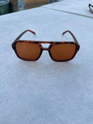 Bruna fyrkantiga solglasögon leopardmönster - Coola solglasögon med fyrkantig form och bruntonade glas. Bågarna har ett trendigt leopardmönster i brunt och svart. Perfekta för att lyfta din stil med en retrovibe. Tillverkade i syntetmaterial. Väldigt snygga och unika till sommaren och hösten när solen är uppe 