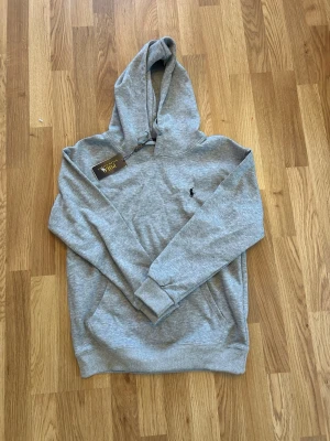 Grå hoodie från Polo Ralph Lauren - Snygg grå hoodie från Polo Ralph Lauren med klassisk huva och känguruficka. Liten broderad logga på bröstet. Perfekt för en avslappnad och stilren look. Mjuk bomullsblandning som känns skön mot huden.