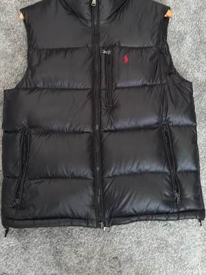 Svart dunväst från Polo Ralph Lauren - Snygg svart dunväst från Polo Ralph Lauren med röd logga på bröstet. Västen har två dragkedjefickor på sidorna och en hög krage. Perfekt för lager-på-lager och håller dig varm med sin vadderade design.