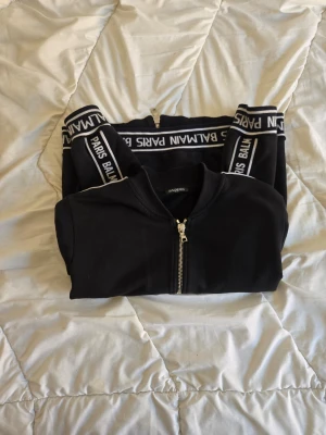 Svart Balmain hoodie med dragkedja - Svart äkta hoodie från Balmain med dragkedja framtill och vita detaljer. Loggan 'PARIS BALMAIN' syns längs ärmar och nederkant. Två fickor med dragkedja och ribbade muddar. Snygg och stilren design som sticker ut.