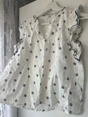 Vit blus med volang och blommönster - Söt vit blus med svarta och ljusblå blommor, volangdetaljer vid ärmhålen och svarta kanter. Blusen är ärmlös och har en lätt, luftig känsla med diskret struktur i tyget. Perfekt för en fräsch och somrig look. Från Lindex, pris kan diskuteras🥰