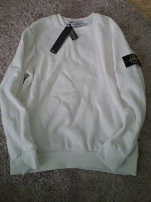 Vit sweatshirt från Stone Island - Säljer en clean vit sweatshirt från Stone Island med den ikoniska svarta patchen med gul och grön logga på ärmen. Tröjan har rund halsringning, ribbade muddar och är tillverkad i mjukt bomullsmaterial. Perfekt för en stilren och avslappnad look. Pris kan diskuteras.