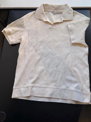 Vit ribbad piké från Zara - Snygg vit piké från Zara med diskret ribbat mönster och klassisk krage. T-shirten har korta ärmar och är tillverkad i mjuk bomull, perfekt för en clean och stilren look.