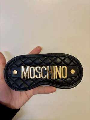Moschino sovmask svart - Snygg svart quiltad sovmask från Moschino med stora guldfärgade bokstäver framtill. Masken har en lyxig känsla med mjukt material och detaljer i guld. Perfekt accessoar för att sova bekvämt och stilfullt. Aldrig använd med saknar förpackning.
