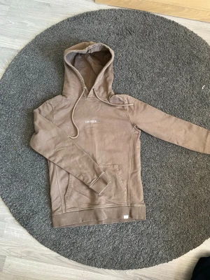 Beige hoodie från Les Deux - Säljer en stilren beige hoodie från Les Deux med broderad logga på bröstet. Hoodien har huva med dragsko och en klassisk känguruficka framtill. Perfekt för en avslappnad och trendig look. 