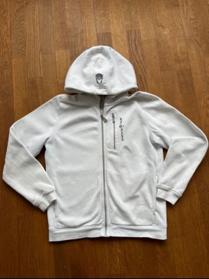 Vit hoodie från Sail Racing - Snygg vit hoodie från Sail Racing med dragkedja framtill och en bröstficka med zip. Hoodien har broderad logga på bröstet och en unik detalj på huvan. Mjuk insida i fleece-liknande material och ribbade muddar vid ärmslut och nederkant.