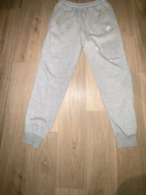 Grå mjukisbyxor från Nike - Gråa mjukisbyxor från Nike med resår i midjan och dragsko. Byxorna har ribbade muddar vid bensluten och en liten vit Nike-logga på vänster lår. Perfekta för en avslappnad stil och tillverkade i mjukt material.