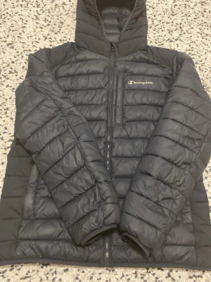 Svart dunjacka från Champion - Snygg svart dunjacka från Champion med huva och dragkedja framtill. Jackan har quiltad design och logga på bröstet. Perfekt för kalla vinterdagar och riktigt skön att bära. Klassisk och sportig stil som funkar till det mesta.