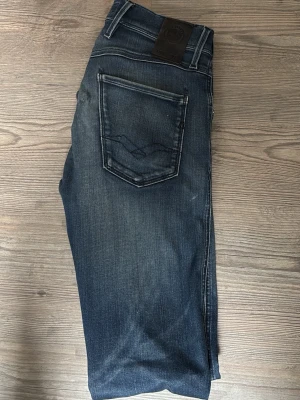 Blå jeans från Replay - Snygga blå jeans från Replay med klassisk femficksmodell och slitna detaljer. Jeansen har dragkedja och knapp i gylfen samt tydliga kontrastsömmar. Perfekta för en avslappnad och trendig look. En liten blå bläck fläck vid vänster ficka som enkelt kan lösas med att göra en liten snygg slitning. 28/32