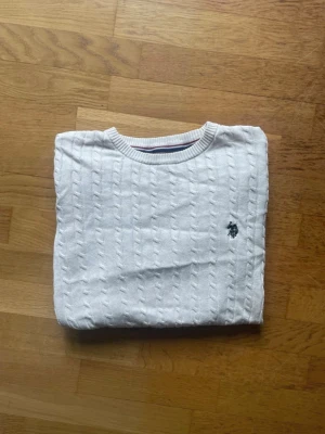 Vit kabelstickad tröja från Polo Ralph Lauren - Kabelstickad vit tröja från Polo Ralph Lauren med rund halsringning och diskret broderad logga på bröstet. Tröjan har långa ärmar och ribbade muddar vid ärmslut och nederkant. Perfekt för lager på lager och klassisk stil.