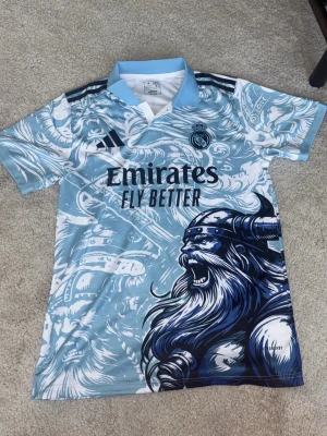 Real Madrid x Adidas vikingatröja - Unik Real Madrid matchtröja i ljusblått och vitt med stort vikingamotiv och detaljerade illustrationer. Tröjan har klubbmärke och sponsortryck på bröstet. Tillverkad i lätt och ventilerande material, perfekt för fotboll eller träning. Skön tröja för alla fotbollsfans!!