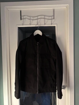 Svart overshirt med dragkedja - Stilren svart overshirt med rak passform och dragkedja framtill. Två stora bröstfickor med lock och klassisk krage. Tillverkad i ett kraftigare tyg som ger en snygg och modern look. Perfekt att bära över en t-shirt för en avslappnad stil.