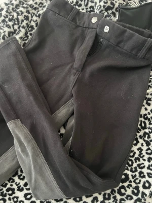 Svarta ridleggings med gråa detaljer - Säljer ett par svarta ridleggings med gråa förstärkningar på insidan av benen. De har hög midja, dragkedja och knapp framtill samt bälteshällor. Perfekta för dig som gillar en sportig och bekväm stil.