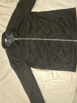 Svart overshirt med dragkedja - Stilren svart overshirt från Weekday med rak passform och två stora bröstfickor med lock. Jackan har en klassisk krage och hel dragkedja framtill. Materialet är slätt och lätt glansigt, vilket ger en modern look. Perfekt att bära som lager över en t-shirt.