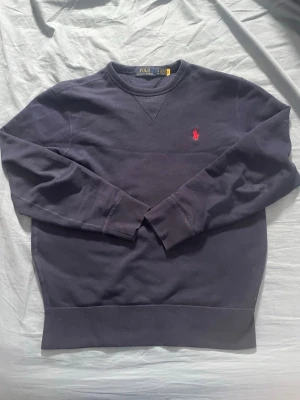 Mörkblå sweatshirt från Polo Ralph Lauren - Mörkblå sweatshirt från Polo Ralph Lauren med klassisk rund halsringning och det ikoniska röda broderade logot på bröstet. Tröjan har långa ärmar och ribbade muddar vid ärmslut och nederkant. Perfekt för en avslappnad och stilren look.