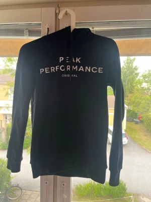 Mörkblå hoodie från Peak Performance - Snygg mörkblå hoodie från Peak Performance med vit text på bröstet. Klassisk modell med huva och dragsnören samt ribbade muddar vid ärmar och nederkant. Perfekt för en avslappnad och sportig stil. Materialet känns mjukt och bekvämt.