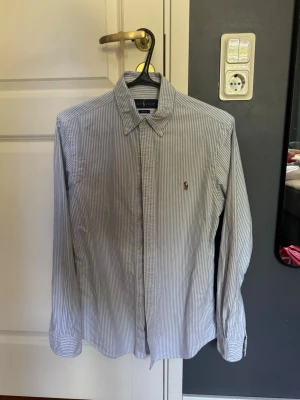 Randig skjorta från Ralph Lauren - Klassisk skjorta från Ralph Lauren med smala blå och vita ränder. Skjortan har lång ärm, knappar framtill och den ikoniska broderade loggan på bröstet. Tillverkad i mjuk bomull och har en tidlös, normal passform.