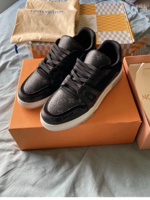 Louis Vuitton svarta sneakers - Snygga svarta Louis Vuitton sneakers med diskret monogrammönster på ovandelen och vita sulor. Skorna har klassisk låg profil, svarta skosnören och detaljer i både mocka och läder. Perfekta för dig som vill ha en exklusiv touch på din outfit.