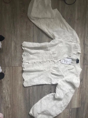 Ballon sleeve blus Gina  - Säljer en vit blus från Gina med broderade blommor och fina spetsdetaljer. Blusen har långärmade puffärmar, knappar framtill och en vågig kant nedtill. Perfekt för dig som gillar romantisk och trendig stil.