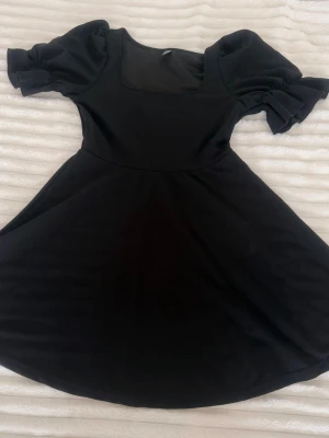 Svart kort puffärmad klänning - Supersnygg svart klänning med puffärmar och rundad urringning. Kjolen har en lätt utställd form som ger en feminin siluett. Perfekt för dig som gillar stilrena och trendiga plagg. Materialet känns mjukt och följsamt.