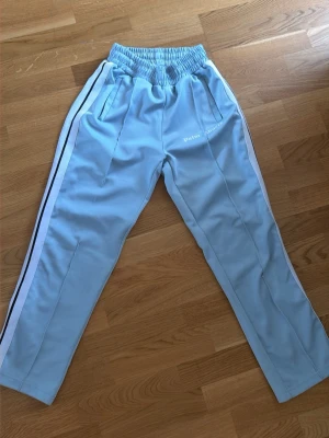 Palm Angels - Säljer ett par ljusblåa mjukisbyxor från Palm Angels med vita sidoränder och elastisk midja. Byxorna har fickor på sidorna och logga tryckt på vänster lår. Perfekta för en chill och trendig streetwear-look.