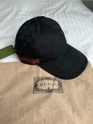 Svart keps  - Snygg svart keps med diskret GG-monogram över hela tyget och klassiska gröna och röda ränder på sidan. Kepsen har böjd skärm och justerbar passform. Materialet känns lyxigt.