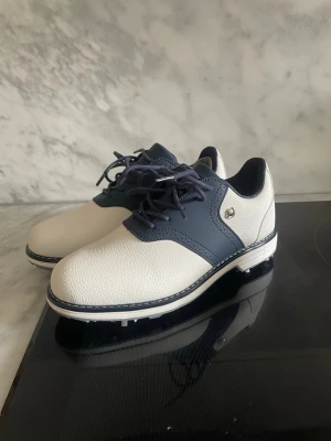 Vita och marinblå golfskor 39 - Snygga golfskor i vitt och marinblått konstläder. Skorna har klassisk snörning, låg profil och detaljerade sömmar. Perfekta för dig som vill ha stilrena och funktionella skor på golfbanan. Storlek 39