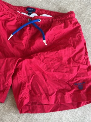 Röda badbyxor från Gant - Snygga röda badbyxor från Gant med blå och vit dragsko i midjan. Klassisk modell med broderad Gant-logga på ena benet. Perfekta för stranden eller poolen, tillverkade i lätt och snabbtorkande material.