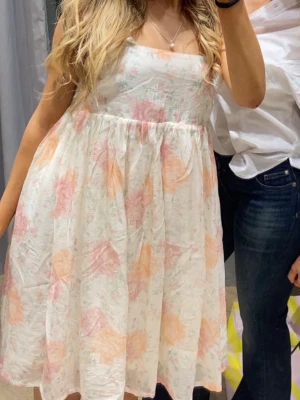 Blommig kort sommarklänning - Söt kort klänning med blommigt mönster i ljusa toner av rosa, orange och vitt. Klänningen har en luftig passform, är ärmlös och har en rak urringning. Perfekt för varma dagar och ger en romantisk vibe.