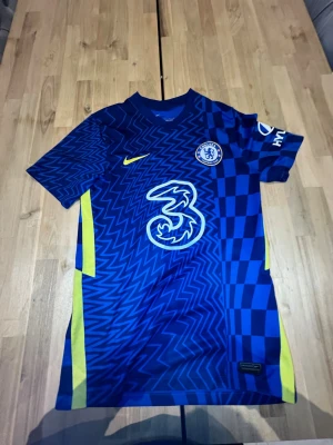 Chelsea FC matchtröja blå Nike - Chelsea FC matchtröja i blått med gula detaljer och zigzag-mönster. Stort vitt '3'-tryck på bröstet, Chelsea-märke och Nike-logga. Tillverkad i lätt och ventilerande polyester, perfekt för fotboll eller träning.