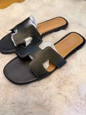 Svarta slip-in sandaler med H-rem - Helt nya Stilrena svarta slip-in sandaler med bred H-formad rem i läderimitation och kontrastsömmar. Dem är aldrig använda. Sandalerna har öppen tå och platt sula med beige innersula för extra komfort. Perfekta för en trendig och avslappnad look. Storlek 42 men passar även mig som har 39 då dem är lite små i storleken.