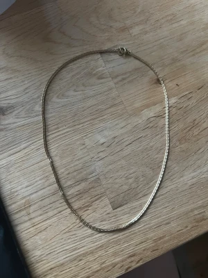 Guldigt halsband  - Snyggt och stilrent halsband i guldfärgad metall från Lily and Rose. Halsbandet har en tunn, boxliknande kedja och en liten rund bricka vid låset med märkets logga. Perfekt för dig som gillar minimalistiska smycken.