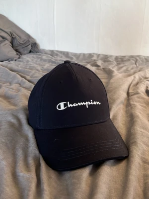 Svart keps från Champion - Snygg svart keps från Champion med klassisk böjd skärm och broderad logga framtill. Justerbar passform med rem baktill. Tillverkad i bomull för en bekväm känsla. Perfekt accessoar för en sportig och avslappnad stil.