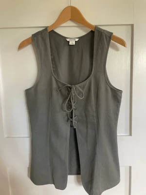 Grå väst med snörning från Monki - Stilren grå väst från Monki i storlek 36. Västen har en djup rundad urringning och snygg snörning framtill som ger en edgy look. Aldrig använd 