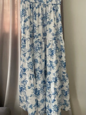Vit och blå blommig långklänning - Långklänning i vitt med blått blommönster och volangdetaljer. Klänningen har en avslappnad passform och ett knytband i midjan för extra form. Perfekt för sommarens alla tillfällen.