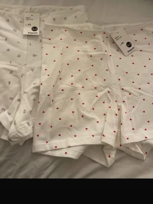 Vita boxerkalsonger med mönster Kappahl - Två par vita sov shorts  från Kappahl i mjuk bomull. Ena paret har små röda hjärtan som mönster, det andra har små blommor i rosa och grönt. Båda har hög midja och elastisk kant upptill. Perfekta för dig som gillar söta detaljer och bekväma plagg 