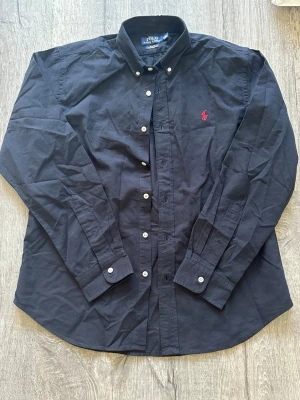 Mörkblå skjorta från Polo Ralph Lauren -  Mörkblå skjorta från Polo Ralph Lauren med röd broderad logga på bröstet. Skjortan har knappar framtill och på kragen samt långa ärmar. Tillverkad i bomull för en skön och stilren look.