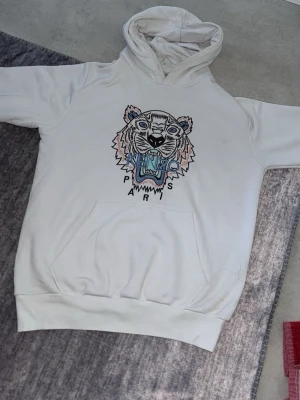 Vit Kenzo hoodie med tigertryck - Säljer en vit hoodie från Kenzo med ikoniskt broderat tigerhuvud och texten 'Paris' på bröstet. Hoodien har huva och stor magficka framtill. Perfekt för dig som vill sticka ut med en snygg streetstyle-look.
