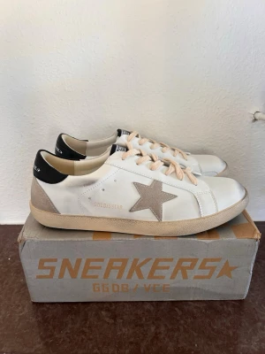 Vita Golden Goose sneakers med stjärna - Snygga vita sneakers från Golden Goose med beige mockadetaljer och ikonisk grå stjärna på sidan. Skorna har svarta detaljer vid hälen och beige snörning. Klassisk låg modell med gummisula och läderöverdel. Perfekta för en trendig och avslappnad look.