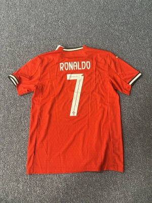 Portugal Ronaldo matchtröja Puma - Röd Portugal matchtröja från Puma med vita detaljer, nummer 7 och Ronaldo på ryggen. Tröjan har korta ärmar, svartvit ribbad krage och ärmslut samt Portugals landslagsemblem på bröstet. Perfekt för dig som älskar fotboll och vill stötta Ronaldo.