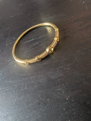 Guldfärgat armband med CC-detaljer - Snyggt guldfärgat cc armband med klassiska CC-loggor längs hela bandet. Armbandet har en stilren och elegant design med små runda detaljer och en fast, rund form. Perfekt för dig som gillar exklusiva accessoarer och vill sticka ut.