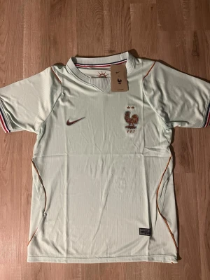 Frankrike VM-2026 Fotbollströja - Snygg ljusgrön fotbollströja med Frankrikes landslagsmärke och två stjärnor på bröstet. Tröjan har korta ärmar, ribbad krage och detaljer i rött, vitt och blått på ärmsluten. Tillverkad i lätt och ventilerande material, perfekt för träning eller match. HELT NY