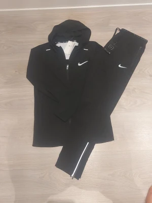 Svart Nike miller tracksiut - Snyggt svart träningsset från Nike med matchande hoodie och byxor. Hoodien har dragkedja och reflekterande detaljer, samt Nike-logga på bröstet. Byxorna har resår i midjan, reflekterande detaljer vid benslut och Nike-logga på benet. Perfekt för löpning eller gym.
