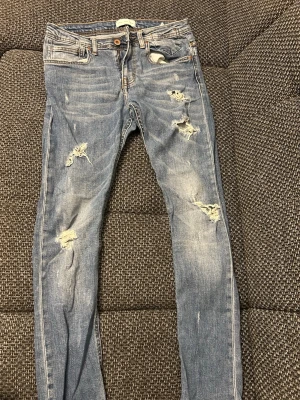 Blå slitna jeans med hål - Säljer ett par blå jeans med slitningar och hål på flera ställen för en cool och avslappnad look. Jeansen har klassisk femficksdesign, knappgylf och smal passform. Perfekta för dig som gillar streetstyle och trendiga detaljer.