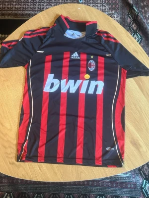 AC Milan matchtröja Maldini #3 - Säljer en klassisk AC Milan matchtröja från Adidas med Maldini och nummer 3 på ryggen. Tröjan är svart och röd med vertikala ränder, klubbmärke och Bwin som sponsor på bröstet. Tillverkad i lätt och ventilerande polyester, perfekt för fotboll eller vardaglig klädsel