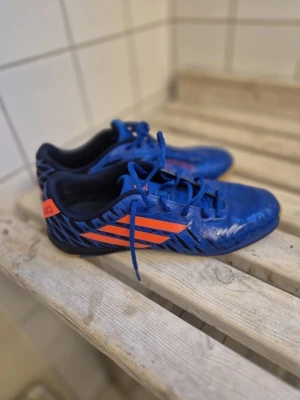 Blå Adidas fotbollsskor inomhus - Säljer ett par blå fotbollsskor från Adidas med orangea detaljer och klassiska ränder på sidan. Skorna har låg profil och är designade för inomhusspel, med texten 'sala' på hälen. Snyggt mönster i svart och blått över hela skon.