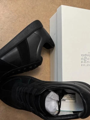 Maison Margiela Gats - Stilrena svarta sneakers från Maison Margiela med ovandel i läder och mocka. Klassisk låg modell med svarta snören och diskret logga på plösen. Snygg och minimalistisk design som passar till det mesta. Helt nya och oanvända! Skön för sommaren😎
