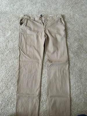Beige chinos från Polo Ralph Lauren - Säljer ett par klassiska beige chinos från Polo Ralph Lauren med rak passform och broderad logga vid fickan. Tillverkade i ett mjukt bomullstyg som känns skönt mot huden. Perfekta för en clean och stilren look.