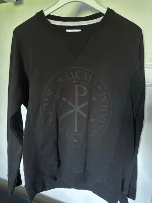 Svart sweatshirt från A.O.CMS XL - Cool svart sweatshirt från A.O.CMS i storlek XL. Tröjan har ett stort, ton-i-ton tryck med grafiskt motiv på bröstet och klassisk rund halsringning. Perfekt för dig som gillar minimalistisk och stilren design.